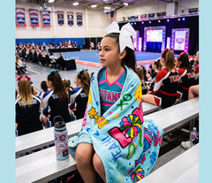 A young cheerleader girl sits on a gym bleachers wrapped in a light blue blanket featuring colorful 'Love 2 Cheer' graphics. She is wearing a large white bow in her hair and a matching cheerleader jersey.