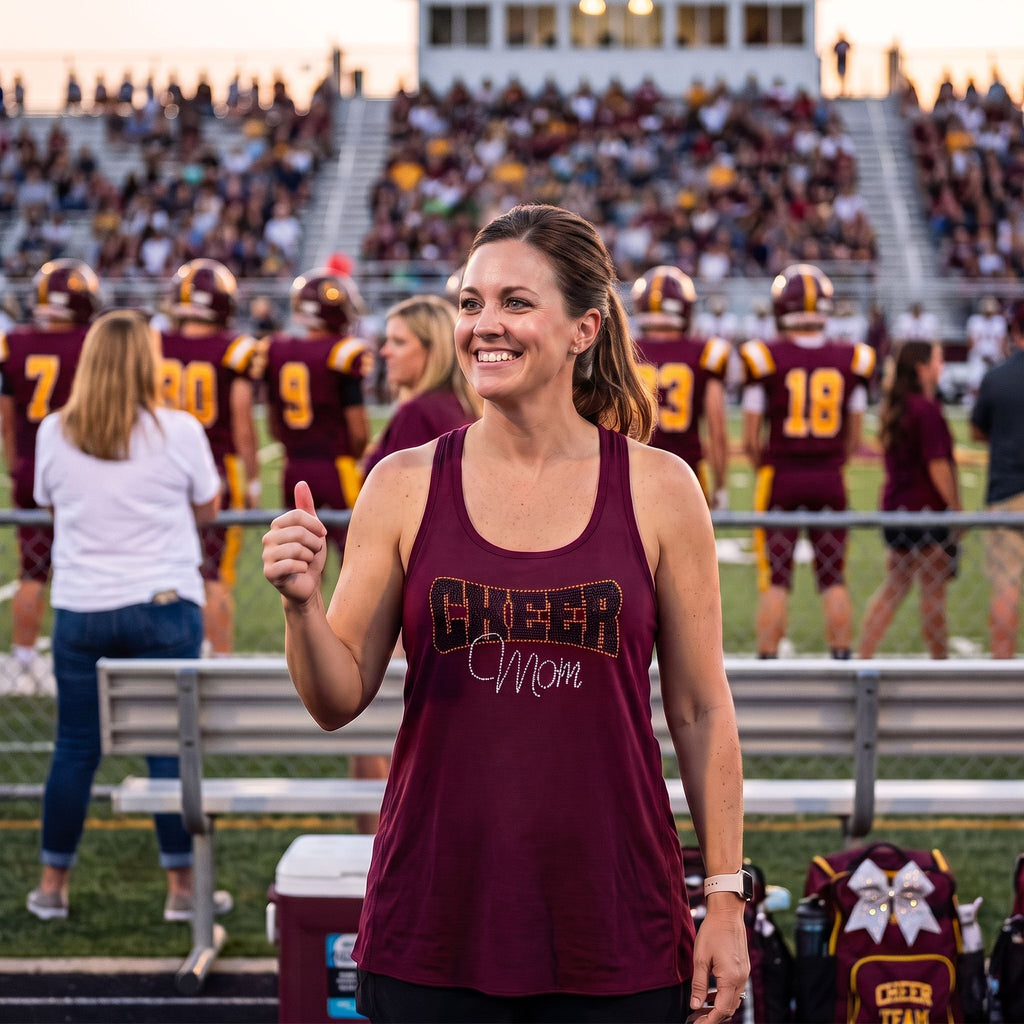 A woman stands on a football field at dusk wearing a maroon tank top with 'CHEER Mom' printed on the front. Cheerleaders and stadium seating are visible in the background.