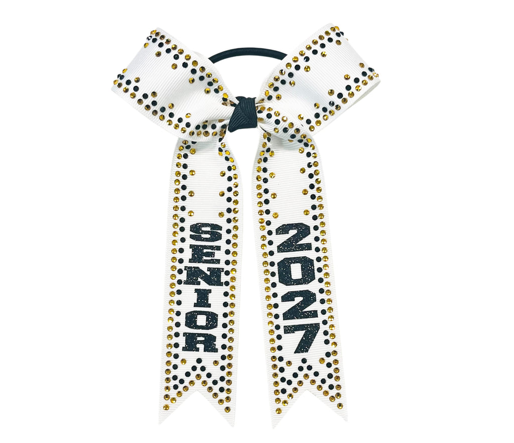 A white cheerleader hair bow with black and gold rhinestone accents, featuring the letters 'SENIOR'  and 2027 in black glitter on one ribbon tail, attached to a black elastic band.
