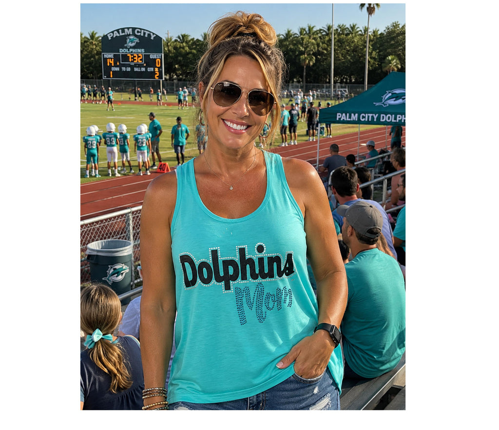 A woman wearing a turquoise tank top with 'Dolphins Mom' printed on the front, standing in a stadium crowd at a football game. The background shows a football field, scoreboard, and other spectators.
