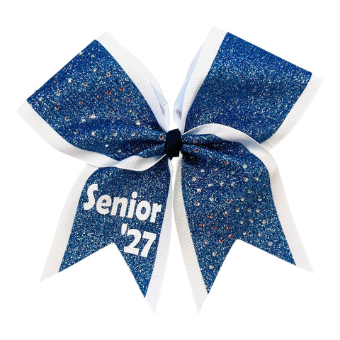 A cheerleader hair bow featuring a columbia blue glitter center with white ribbon borders and white lettering that reads 'Senior '27' on the lower left ribbon, shown on a plain white background.