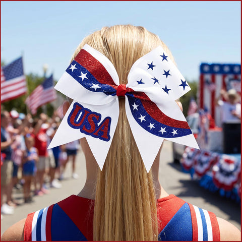 A blonde girl seen from behind wearing a large patriotic cheerleader hair bow with white panels, red glitter stripes, blue star sections, and 'USA' lettering, at an outdoor parade or festival.