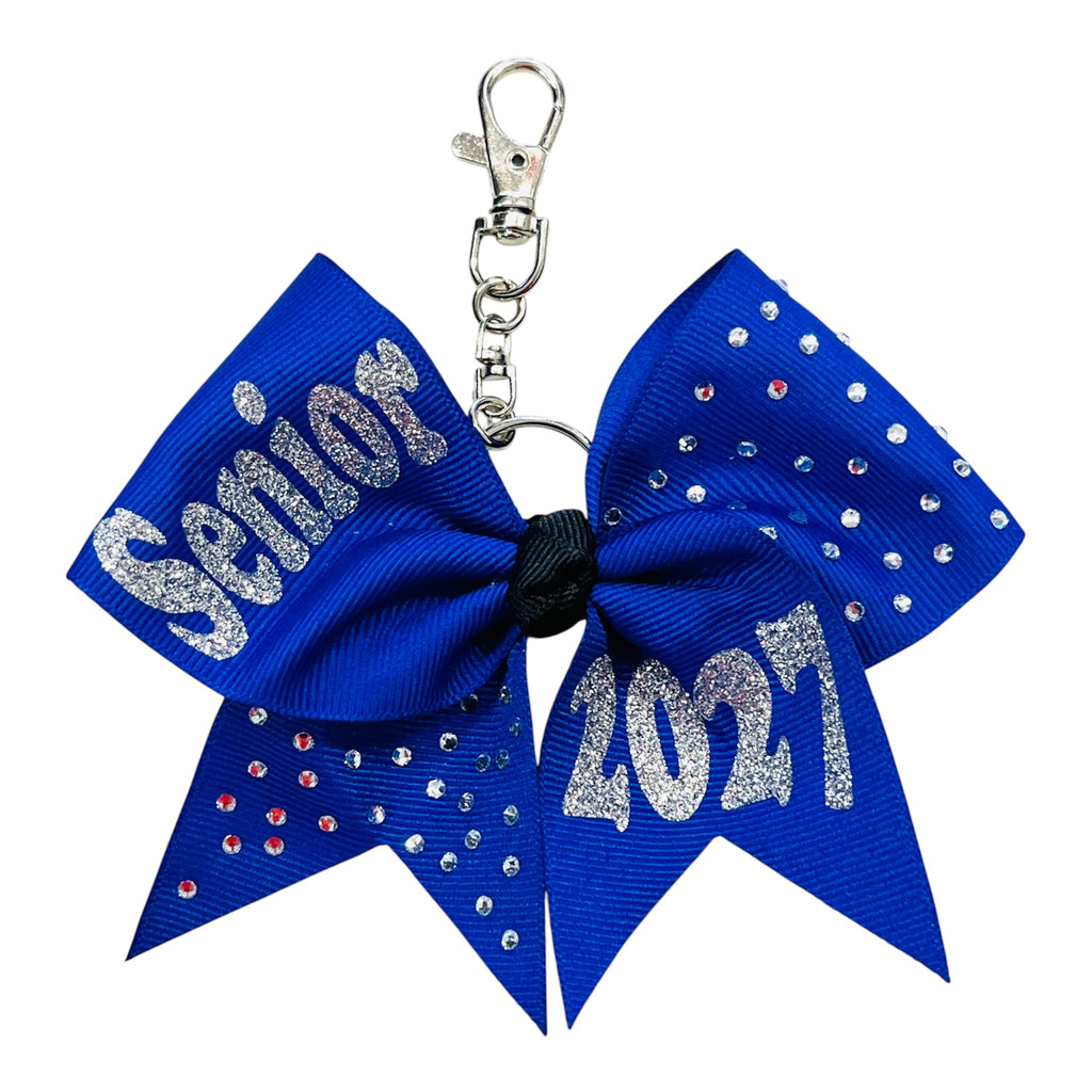 A royal blue cheerleader-style keychain bow with silver glitter text reading 'Senior 2027' and rhinestone embellishments, attached to a silver clasp on a white background.