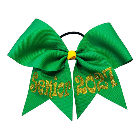 A green cheerleader hair bow with 'Senior 2027' written in yellow gold glitter on the tails, attached to a black elastic band with a yellow gold center knot, shown on a plain white background.