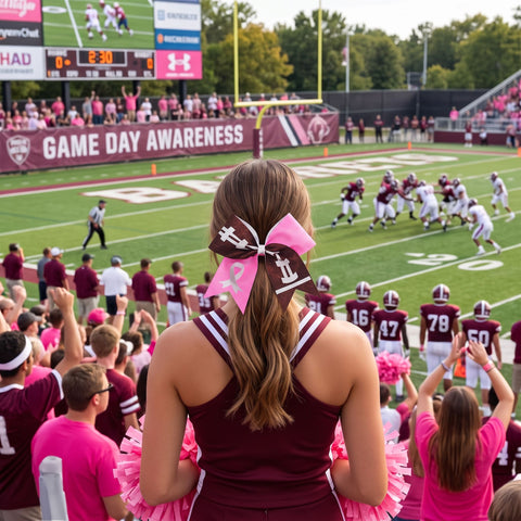 A cheerleader in a maroon and pink uniform stands on a football field, seen from behind, with a large pink, football and white hair bow in her hair. Other cheerleaders and fans in pink and maroon cheer around players on the field during a game.